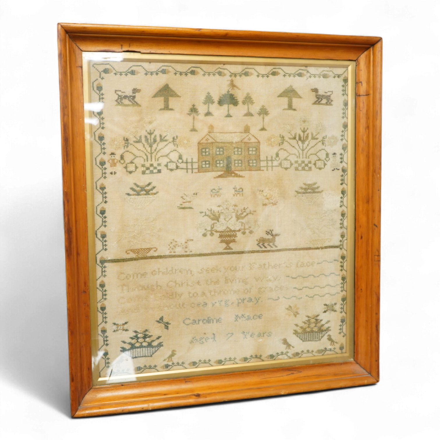 A framed 19th century sampler by Caroline Mace aged 7 years. Worked in cross stitch with the top half showing a scene of a house and trees, the lower half with a verse and various spot motifs within a vining border. 48.5